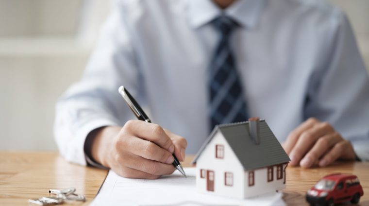 Signing Real Estate Contract for Property and Vehicle Purchase A businessman signing a real estate contract with house and car models on the table, representing property and vehicle purchase agreements.